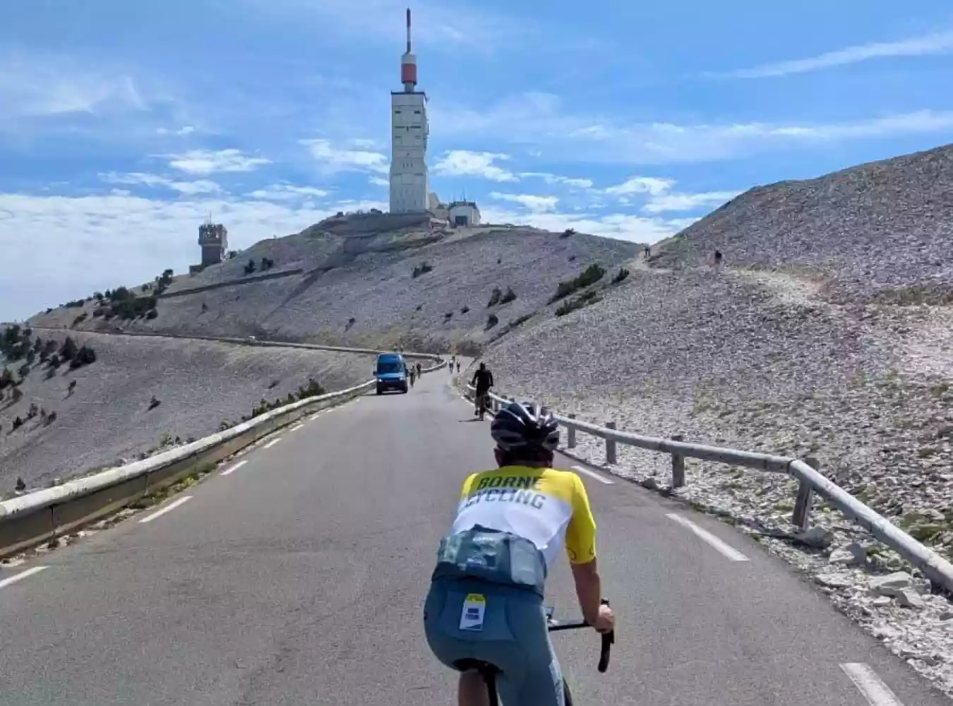 A cyclist in yellow white and green kit riding up towards the summit of Mont Ventoux. The communications tower is prominent in the distance. The landscape is rocky and barren.