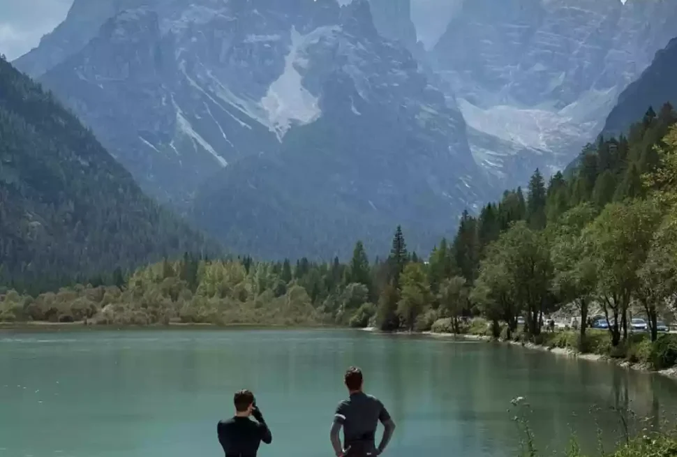 2 people in cycling clothes standing by a lake looking up at the Dolomites.