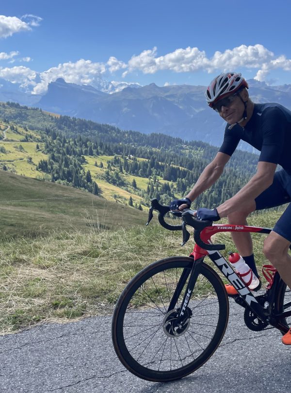 A cyclist at the top of a mountain road with a view of the Mont Blanc range behind him.