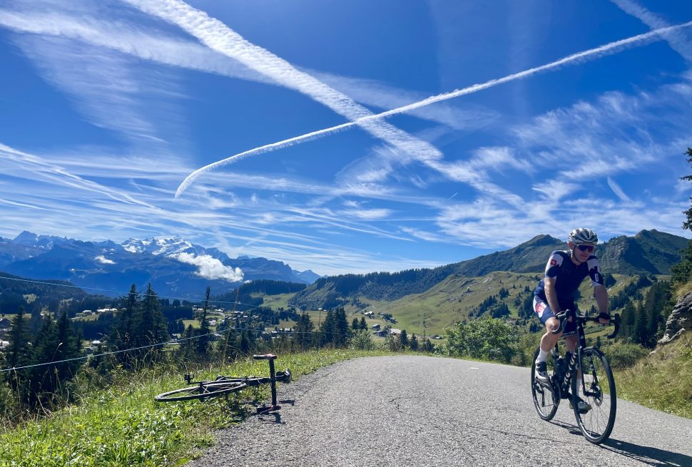 A cyclist in blue kit on a blue sky day with contrails criss crossing the sky. The Mont Blanc is in the background.
